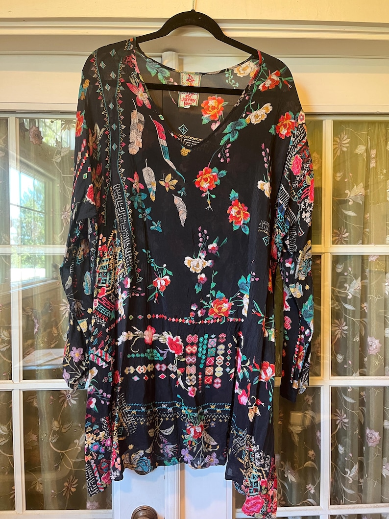 May include: A black long-sleeve tunic featuring a vibrant floral print with red, pink, and teal accents. The garment has a V-neckline and a label at the inner collar that reads Johnny Was. The fabric displays a mix of botanical and geometric patterns.