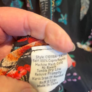 May include: A black garment with a colorful floral pattern and a white care label that reads: Style: C10118A-9X, Self: 100% Cupra Rayon, Machine Wash Cold, No Bleach, Tumble Dry Low, Remove Promptly, Warm Iron If Needed, Rn# 87291, Made In China.