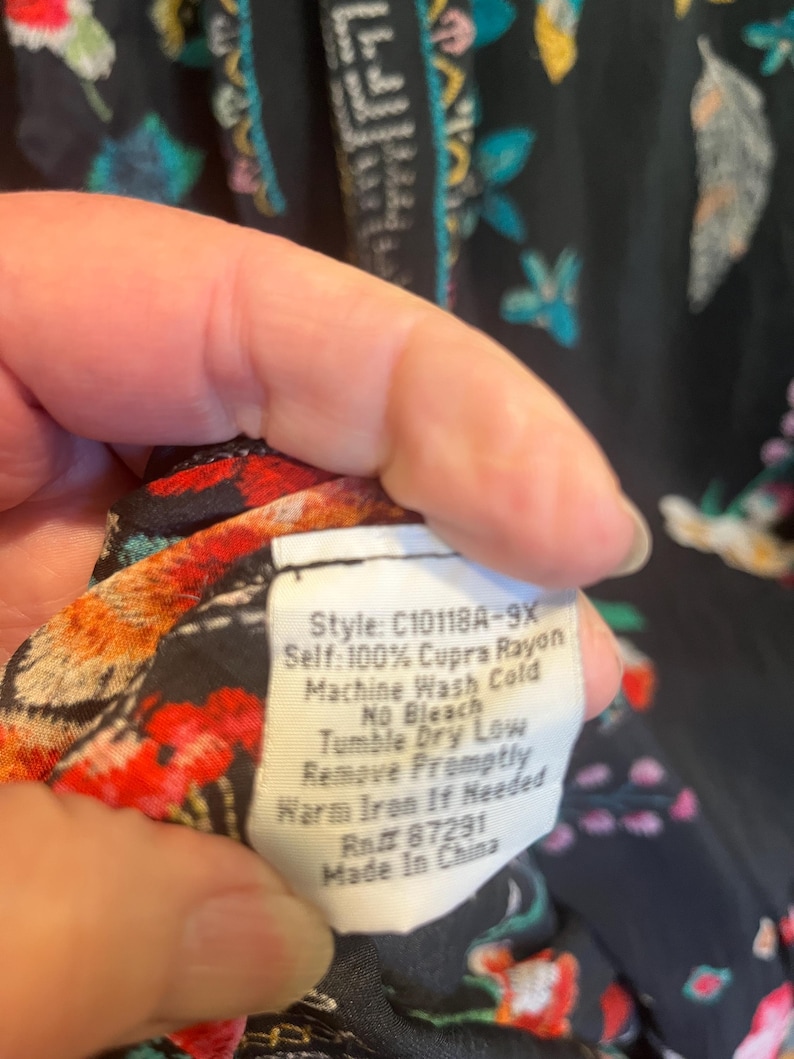 May include: A black garment with a colorful floral pattern and a white care label that reads: Style: C10118A-9X, Self: 100% Cupra Rayon, Machine Wash Cold, No Bleach, Tumble Dry Low, Remove Promptly, Warm Iron If Needed, Rn# 87291, Made In China.