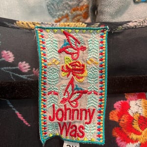 May include: A black garment featuring a rectangular clothing label with the text Johnny Was in red embroidery. The label is decorated with colorful embroidered floral and butterfly motifs in red, yellow, and blue, and includes a white size tag reading 2X.
