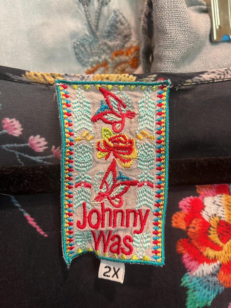 May include: A black garment featuring a rectangular clothing label with the text Johnny Was in red embroidery. The label is decorated with colorful embroidered floral and butterfly motifs in red, yellow, and blue, and includes a white size tag reading 2X.