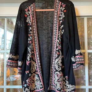 May include: A black open-front cardigan featuring intricate embroidery along the front panels and sleeve cuffs. The embroidery includes floral and leaf patterns in shades of pink, white, and light blue, with geometric borders along the edges.