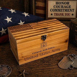 May include: A wooden box with an engraved American flag design on the lid. The box is personalized with the text "Corporal: Josh Bohen United States Army." An American flag, dog tags, and a commemorative coin are also visible.