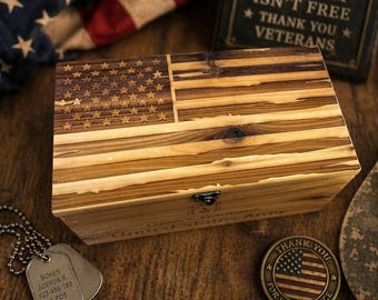 May include: Wooden box with an engraved American flag design. The box is made of light-colored wood and has a hinged lid. Also visible are military dog tags, a commemorative coin, and a sign that reads "Freedom Isn't Free, Thank You Veterans."