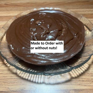 May include: A round, clear glass serving dish filled with a thick, dark brown dessert. The dessert has a glossy surface and appears to be a chocolate pudding or mousse. Text on the image reads: Made to Order with or without nuts!
