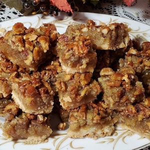 May include: A plate of pecan squares, a type of dessert bar, with a light brown crust and a pecan topping. The squares are arranged in a pile on a white plate with a gold floral pattern.