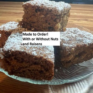 May include: A clear glass plate holds several square slices of cake, dusted with powdered sugar. The cake is light brown with visible nuts and raisins. Text on the image reads "Made to Order! With or Without Nuts and Raisins."