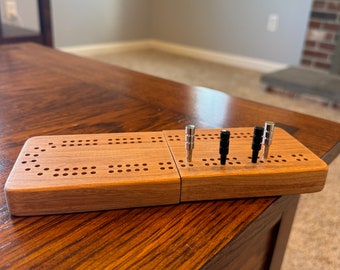Peut inclure: Un jeu de cribbage en bois avec une finition brun clair. Le plateau est fermé et des chevilles sont insérées dans les trous.