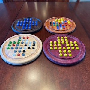 May include: Four wooden game boards with different colored marbles. The boards are made of different types of wood, including walnut, maple, and cherry. The marbles are arranged in a pattern on the boards.