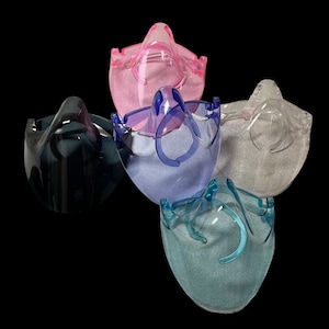 Op de afbeelding: Vijf transparante gezichtsbeschermers van plastic in verschillende kleuren: roze, blauw, grijs, zwart en teal. De schermen zijn ontworpen om het gezicht en de ogen van de drager te beschermen.