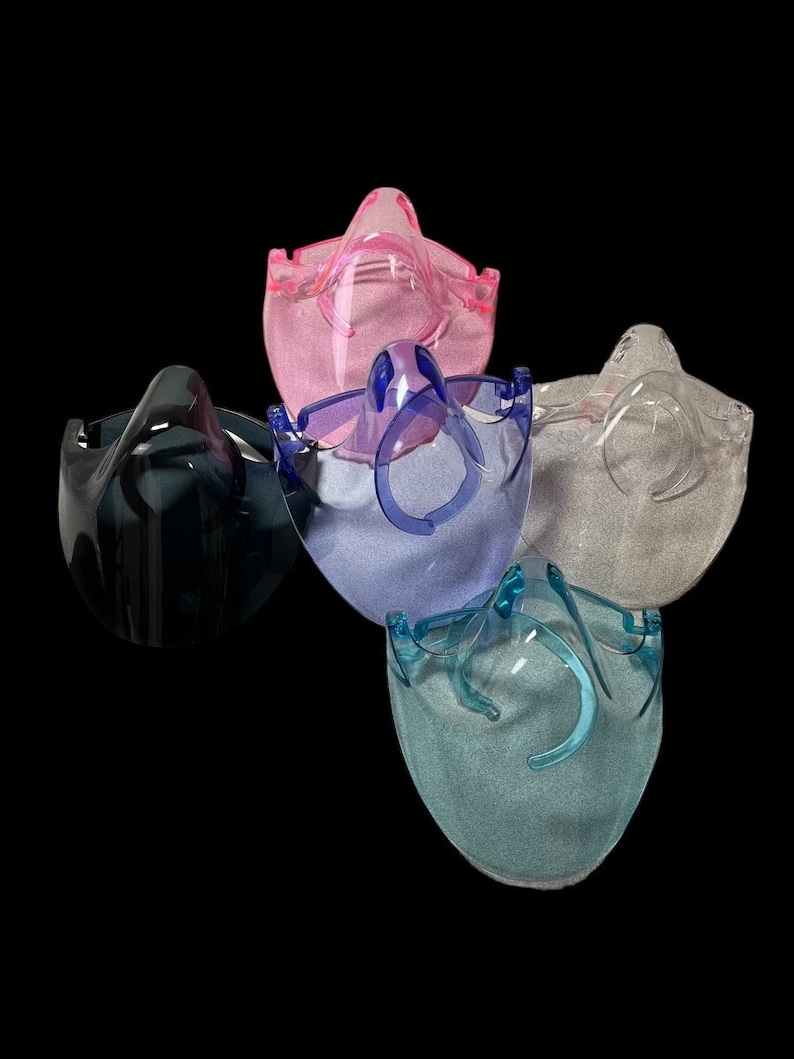 Op de afbeelding: Vijf transparante gezichtsbeschermers van plastic in verschillende kleuren: roze, blauw, grijs, zwart en teal. De schermen zijn ontworpen om het gezicht en de ogen van de drager te beschermen.
