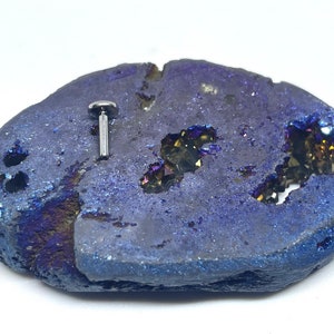 May include: A silver barbell piercing with a small clear gem on the top. The barbell is on a blue and purple geode.