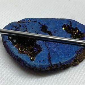 May include: A polished, silver-colored metal rod rests on a vibrant blue geode with sparkling, golden-brown crystal formations. The geode's surface has a textured appearance, creating a striking contrast with the smooth rod.