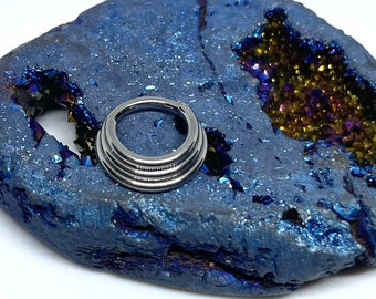 May include: A silver, multi-layered, seamless ring for body piercing. The ring is displayed on a blue and purple geode.