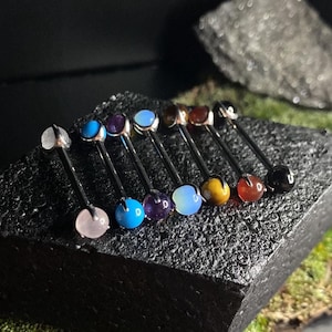May include: A collection of body jewelry, each with a different colored gemstone. The jewelry is displayed on a textured black surface, with a dark background. The gemstones include pink, blue, purple, and brown. The jewelry is made of silver metal.