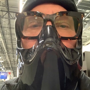 Op de afbeelding: Een persoon draagt ​​een zwart gezichtsmasker met een helder vizier en schildpadbril. De persoon draagt ​​een zwarte beanie en een zwart jack.