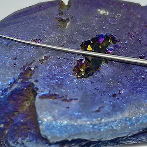 May include: A thin, silver, needle-like tool on a purple and gold iridescent agate geode.