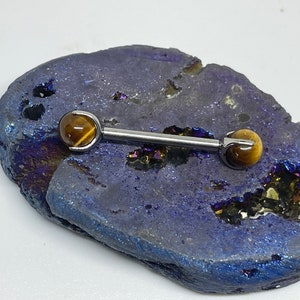 May include: A silver-colored barbell-style body piercing with tiger's eye gemstone ends. The jewelry is displayed on a blue and purple geode.