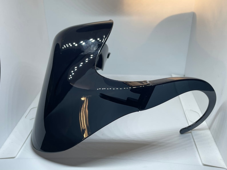 Op de afbeelding: Een zwart plastic gezichtsbeschermer met een gebogen ontwerp en een enkele band die over het hoofd gaat.