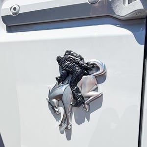 May include: A silver-colored metal emblem featuring a bucking horse with a black Bigfoot figure riding on its back, mounted on the white rear panel of a vehicle next to a black spare tire and a red taillight assembly.