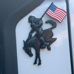 May include: Black emblem of a figure riding a horse, holding a flag with the American flag design. The emblem is mounted on a white surface, with a tire visible on the left side.