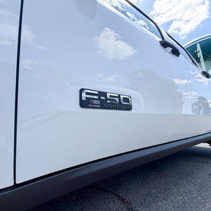 May include: A white Ford F-150 Light Duty truck. The truck's side panel features a chrome emblem with the text "F-150 LIGHT DUTY". The image is taken from a low angle, showing the wheel and the sky.