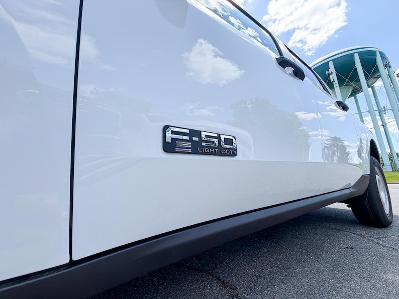 May include: A white Ford F-150 Light Duty truck. The truck's side panel features a chrome emblem with the text "F-150 LIGHT DUTY". The image is taken from a low angle, showing the wheel and the sky.
