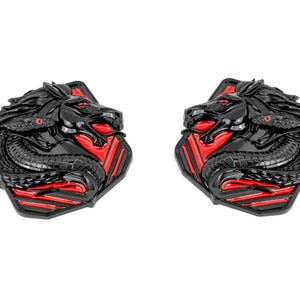 May include: A pair of black metal decorative emblems with a red and black geometric design. Each emblem features a stylized black horse head with a snake wrapped around its neck.