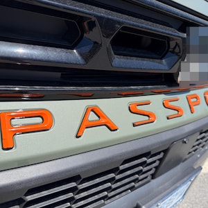 May include: The front grille of a vehicle featuring the word PASSPORT in bold orange lettering. The vehicle body is a muted sage green color, contrasted by a glossy black upper grille section with rectangular openings and a black lower bumper.