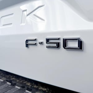 May include: Close-up of a white vehicle's rear with the chrome F-150 emblem prominently displayed. The letters are outlined in black, creating a contrast against the white surface. The word 'FICK' is also visible.