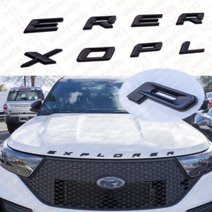 May include: Black car emblem letters spelling "EXPLORER". The letters are displayed individually and also on the hood of a white SUV. The emblem letters are made of a glossy black material.
