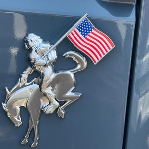 May include: A chrome emblem featuring a rider on a horse, holding a US flag. The emblem is mounted on a blue surface. The flag has red and white stripes and a blue field with white stars.