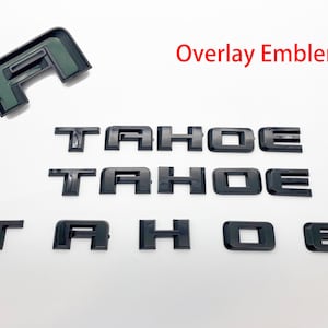 May include: Black overlay emblems for a Tahoe vehicle. The emblems are in the shape of the letters "TAHOE" and are arranged in three rows. The top row has the letters "TAHOE" in a larger font size. The middle row has the letters "TAHOE" in a smaller font size. The bottom row has the letters "TAHOE" in the smallest font size. The emblems are on a white background. Overlay Emblems is written in red text at the top of the image.