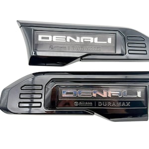 May include: Two glossy black automotive side emblems featuring the text DENALI in bold silver lettering. Below the main text, the emblems display the Allison Transmission logo and the word DURAMAX. Each piece includes three horizontal vent-like slots.