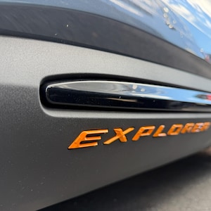 May include: A matte grey vehicle side panel featuring a glossy black horizontal trim piece and the word EXPLORER embossed in bright orange lettering. The surface has a textured finish, and the design includes a sleek, modern aesthetic detail.