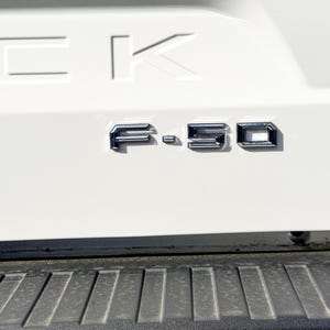 May include: Close-up of a white truck bed with chrome lettering. The letters "F-150" are prominently displayed, with the "CK" logo above. The lower portion of the image shows a black textured bumper.