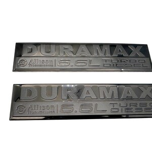 May include: Two black Duramax 6.6L Turbo Diesel emblems. Each emblem features the word DURAMAX in raised lettering, with Allison Transmission and 6.6L Turbo Diesel text below. The emblems are rectangular with a glossy finish.