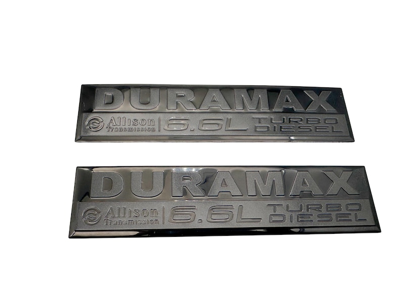 May include: Two black Duramax 6.6L Turbo Diesel emblems. Each emblem features the word DURAMAX in raised lettering, with Allison Transmission and 6.6L Turbo Diesel text below. The emblems are rectangular with a glossy finish.