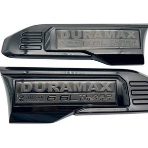 May include: Two black Duramax 6.6L Turbo Diesel emblems. Each emblem features the word DURAMAX, with Allison Transmission and 6.6L Turbo Diesel text below. The emblems are black with a glossy finish.