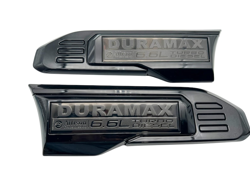 May include: Two black Duramax 6.6L Turbo Diesel emblems. Each emblem features the word DURAMAX, with Allison Transmission and 6.6L Turbo Diesel text below. The emblems are black with a glossy finish.