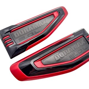 May include: Two automotive engine emblems featuring a red and black finish with the embossed text DURAMAX 3.0L. These molded plastic components have a sleek, angular design with three horizontal red slats on one end and a textured center panel.