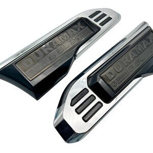 May include: Two chrome and black Duramax 6.6L Turbo Diesel emblems. The emblems have a rectangular shape with the text "DURAMAX" and "6.6L Turbo Diesel" in a dark grey colour. The emblems are for a vehicle.