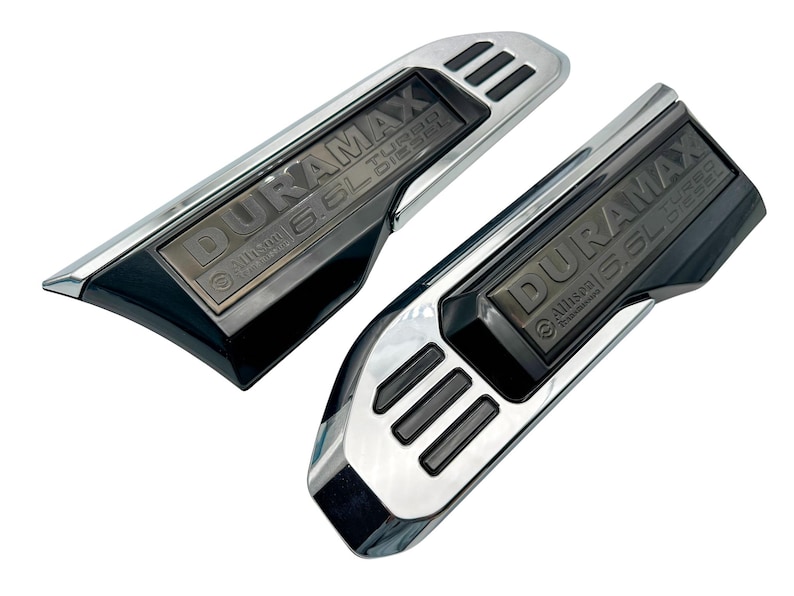 May include: Two chrome and black Duramax 6.6L Turbo Diesel emblems. The emblems have a rectangular shape with the text "DURAMAX" and "6.6L Turbo Diesel" in a dark grey colour. The emblems are for a vehicle.