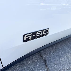 May include: A black and silver F-150 Light Duty emblem with a Canadian maple leaf on a white vehicle. The emblem is rectangular and mounted on the side of the vehicle.