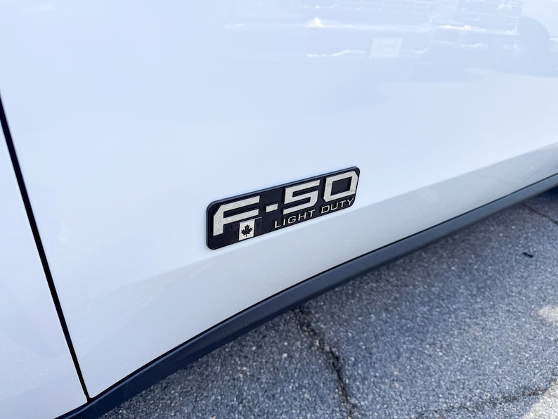 May include: A black and silver F-150 Light Duty emblem with a Canadian maple leaf on a white vehicle. The emblem is rectangular and mounted on the side of the vehicle.