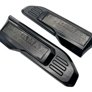 May include: Two black Duramax 6.6L Turbo Diesel engine covers. The covers are angled and have the Duramax logo and Allison 6.6L Turbo Diesel text embossed on them. The covers have a glossy finish.