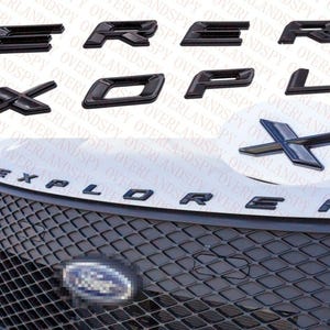 May include: Black "Explorer" emblem letters and an "X" emblem. The image shows individual letters and the complete word "EXPLORER" on a vehicle's hood and grill. The emblems are glossy and appear to be made of plastic.