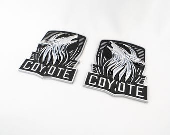 May include: Two black and silver metal car emblems with a stylized coyote design and the word "COYOTE" in white lettering.