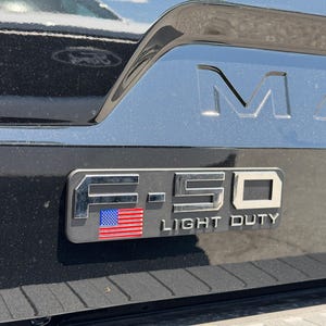 May include: Close-up of a black truck's tailgate with the embossed word "MAX" and a chrome emblem. The emblem reads "F-150 LIGHT DUTY" with an American flag design.