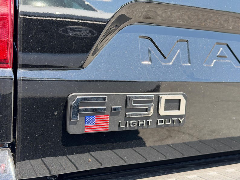 May include: Close-up of a black truck's tailgate with the embossed word "MAX" and a chrome emblem. The emblem reads "F-150 LIGHT DUTY" with an American flag design.